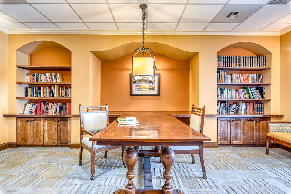 A cozy reading or study area with a wooden table and two chairs in front of built-in bookshelves filled with books. The walls are painted in warm tones with an orange accent wall in the center alcove. A hanging light fixture is above the table, and there is a framed picture on the accent wall. The floor is carpeted with a patterned design.