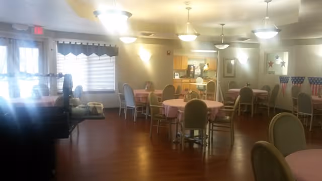 A dining room with multiple round tables covered with pink tablecloths and surrounded by chairs. The room has wooden flooring, ceiling lights, and a small kitchen area in the back. There are windows with blinds and patriotic decorations on the walls.