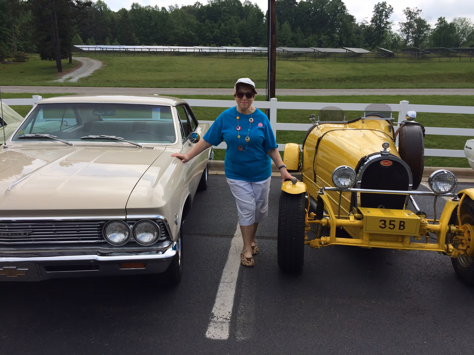 An elderly woman wearing a blue shirt, white cap, white pants, and sunglasses stands between two vintage cars parked in an outdoor lot. She is touching both cars, a beige classic car on her left and a yellow vintage car on her right. Behind her is a white fence and a grassy area with trees and solar panels in the distance.