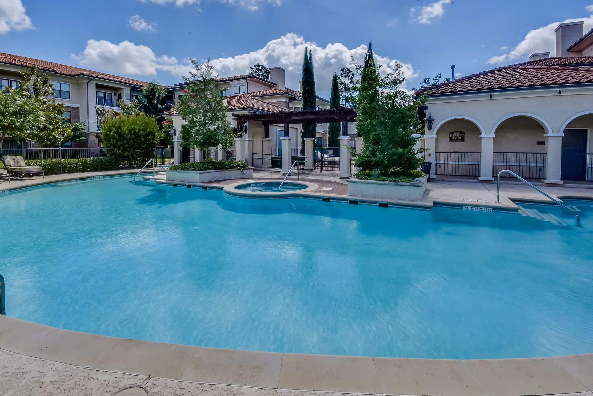 Outdoor swimming pool area at a senior living facility with clear blue water, surrounded by lounge chairs, greenery, and Mediterranean-style buildings with tiled roofs under a partly cloudy sky.