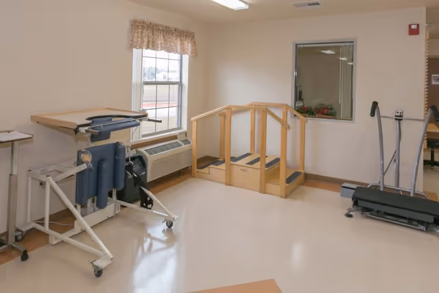 A rehabilitation room with physical therapy equipment including a standing frame, a small set of wooden stairs with handrails, and a treadmill. The room has a window with a valance, beige walls, and a polished floor.