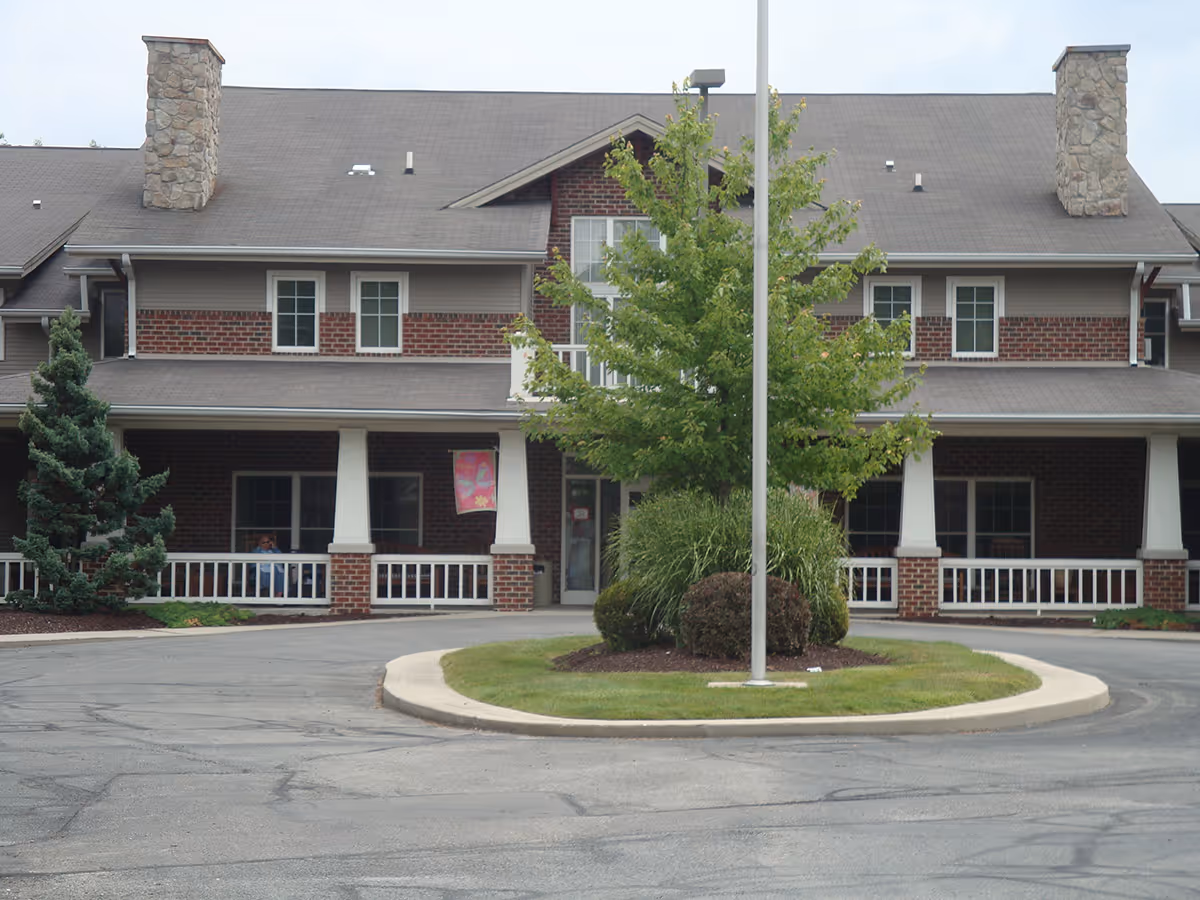 Front exterior view of a two-story brick building with a covered porch, white railings, and two stone chimneys. There is a circular driveway with a landscaped island featuring a tree and bushes in front of the building.