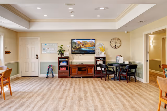 A cozy common area in a senior living facility featuring a wall-mounted flat-screen TV displaying a scenic waterfall image, a wooden media console with shelves holding various items, a round table with four chairs, a wall clock, and a door to the left. The room has beige walls with a green lower half, wood trim, and wood-look flooring.