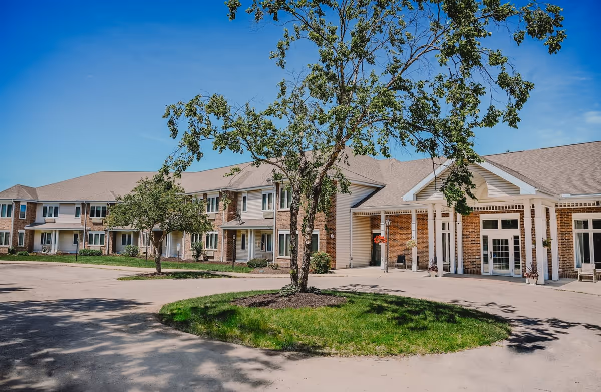 Exterior view of a senior living facility building with a circular driveway and a tree in the center. The building has two stories with multiple windows and a covered entrance with white pillars. The sky is clear and blue.