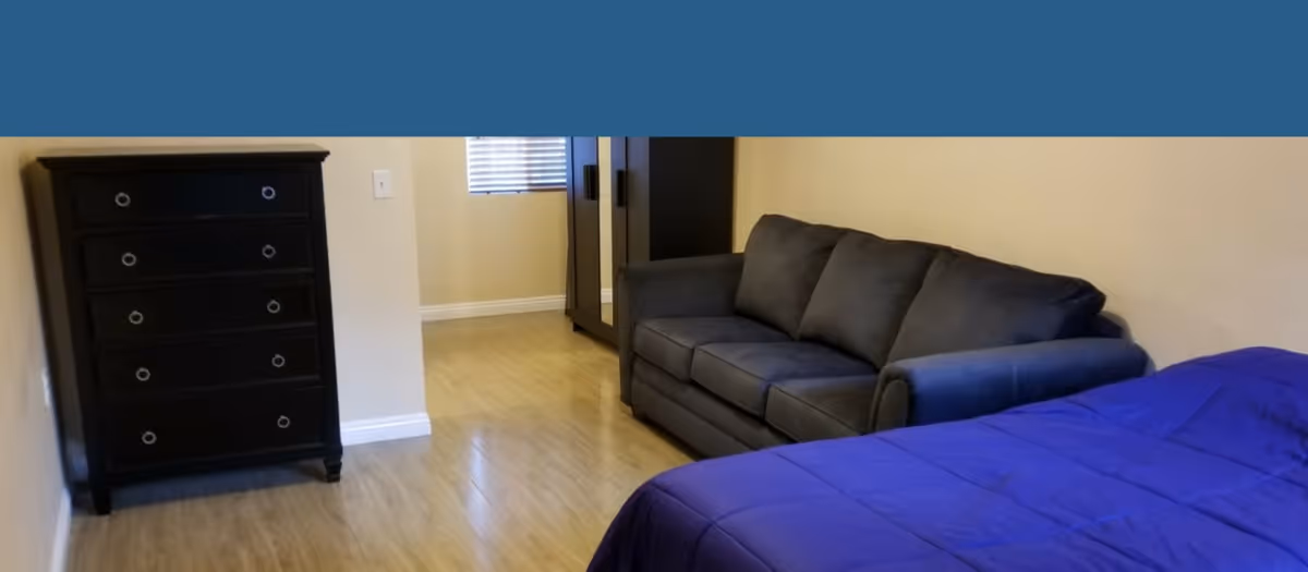 Interior view of a room with a wooden floor, a black chest of drawers on the left, a dark gray sofa against the right wall, and a bed with a blue comforter in the foreground. A window with blinds and a wardrobe are visible in the background.