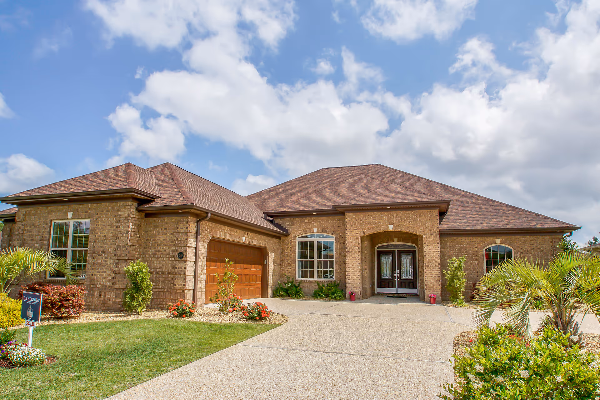 Exterior view of a single-story brick house with a brown shingled roof, a double garage door, and a driveway. The front yard is landscaped with green grass, small bushes, and palm plants under a partly cloudy blue sky.