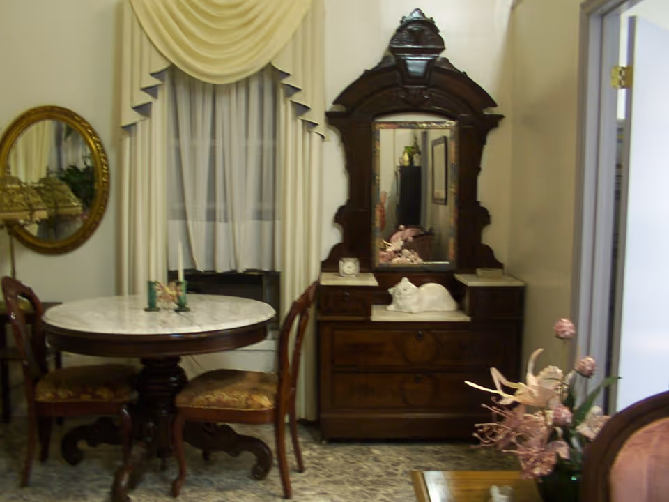 A vintage-style interior room featuring a round marble-top table with four wooden chairs, a large ornate wooden dresser with a mirror, a white ceramic cat figurine on the dresser, floral arrangements, and a window with cream-colored drapes and sheer curtains.