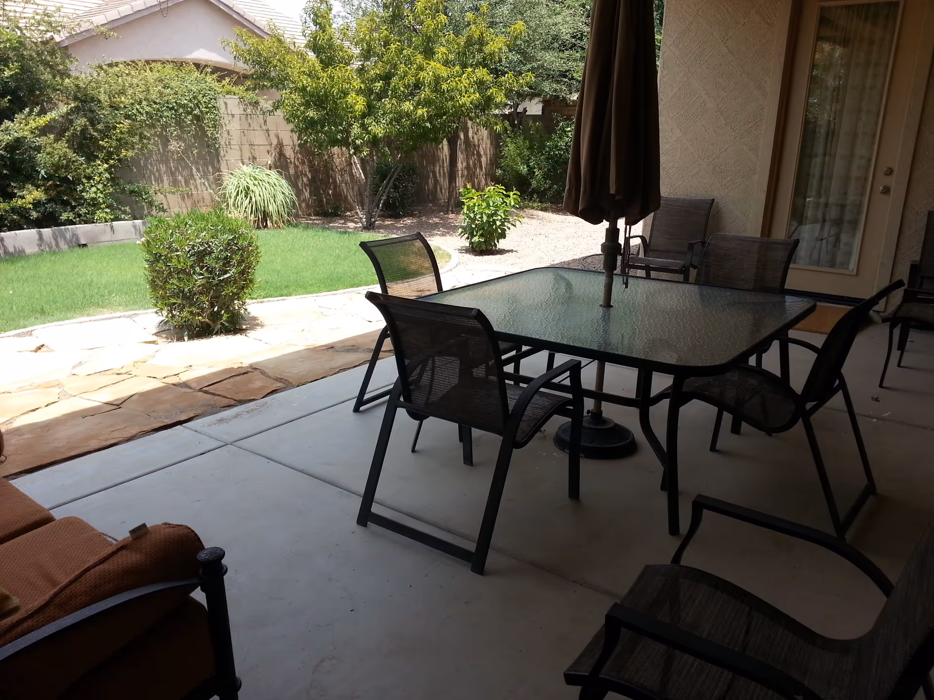 Outdoor patio area with a glass-top table and six mesh chairs, an umbrella in the center of the table, and a cushioned chair on the left. The patio overlooks a garden with green grass, bushes, trees, and a stone pathway.