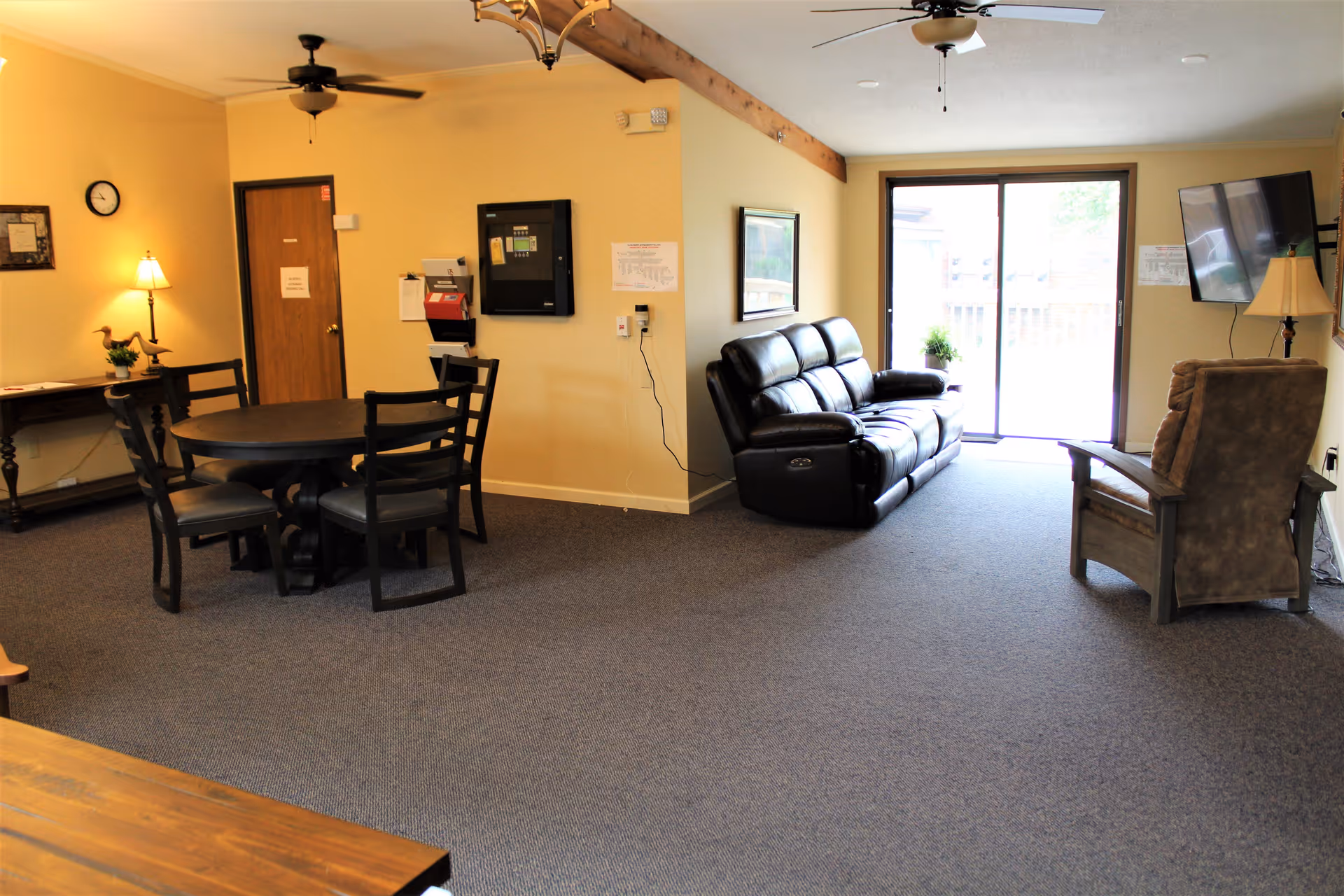 A cozy living room area in Claremont Retirement Village featuring a black leather reclining sofa, a brown upholstered armchair, a wall-mounted flat-screen TV, and a sliding glass door leading outside. There is a round dining table with four chairs, a small wooden side table with a lamp and decorative items, and ceiling fans with lights. The walls are painted light yellow and the floor is carpeted.