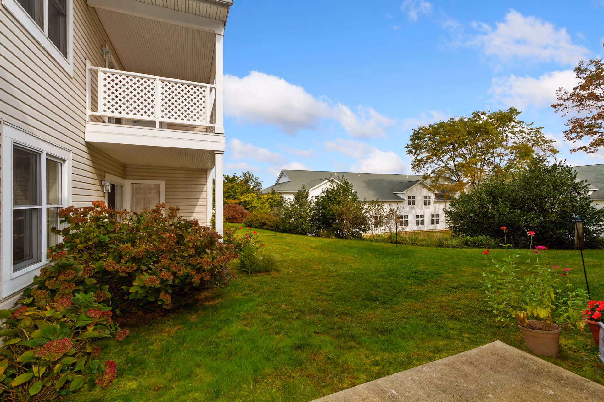 View of a green lawn with bushes and potted plants next to a beige building with white trim and a balcony. In the background, there is another white building with a green roof surrounded by trees under a partly cloudy blue sky.