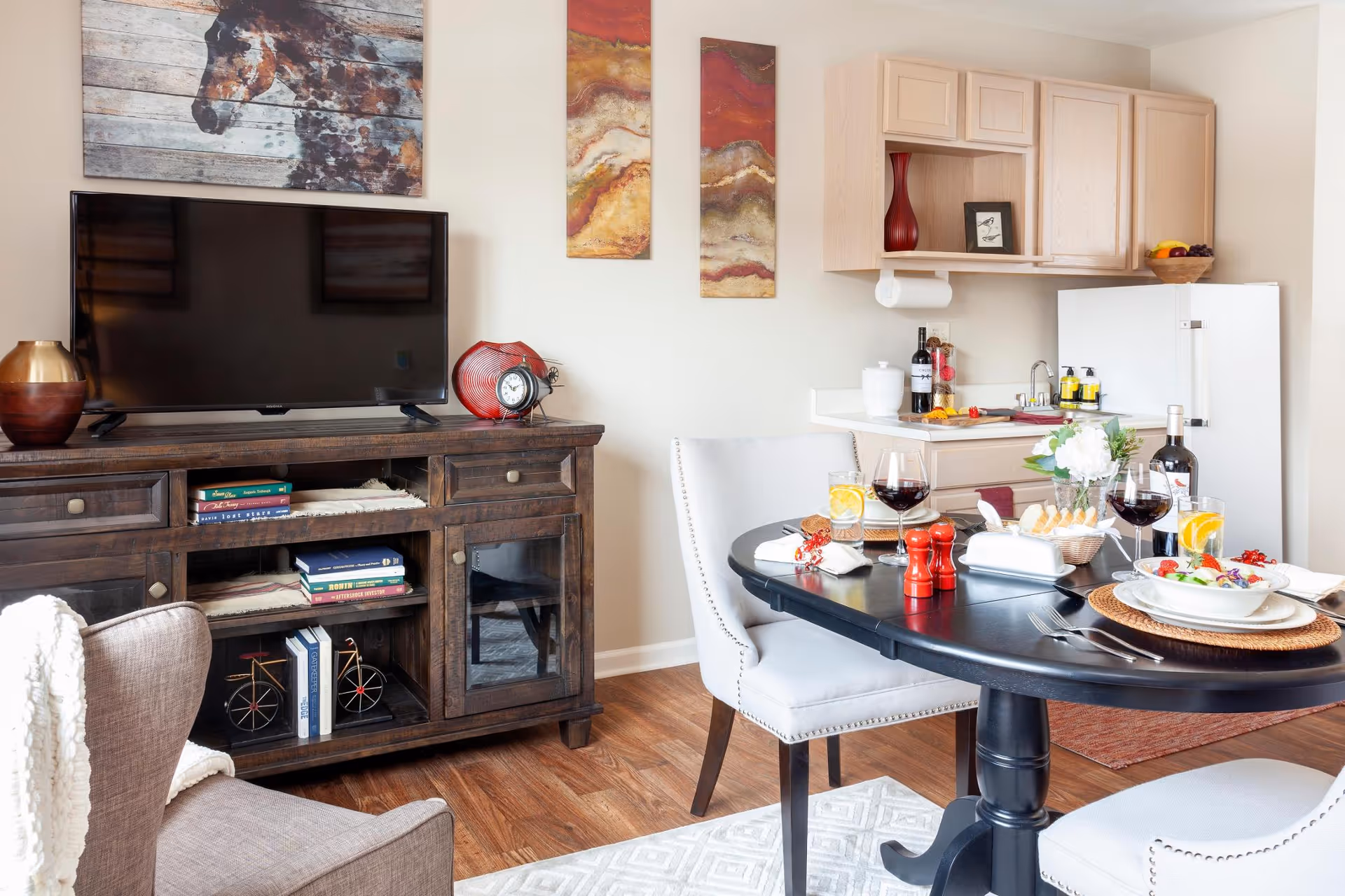 A cozy living and dining area featuring a dark wooden TV stand with a flat-screen TV, decorative items, and books. Adjacent to it is a small kitchen area with light wood cabinets, a white refrigerator, and a countertop with a sink and various items. In the foreground, a round black dining table is set with plates of food, glasses of wine and water, and decorative flowers. The room has wooden flooring and light-colored walls adorned with artwork.