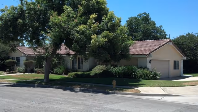 Single-story residential building with a tiled roof, surrounded by green trees and bushes, situated on a corner lot with a sidewalk and street in front.