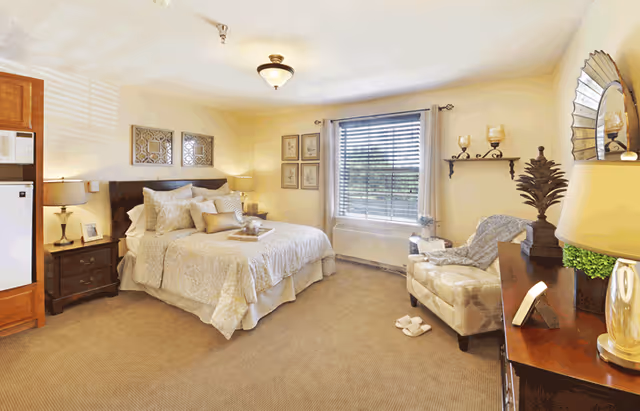 Bright, well-decorated bedroom with a made double bed, nightstands, an armchair, dresser, and a window with blinds.