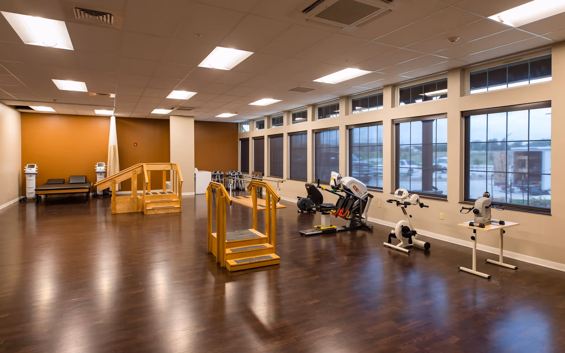 A spacious rehabilitation or physical therapy room with wooden handrails and steps for mobility exercises, exercise bikes, and other physical therapy equipment. The room has large windows along one wall letting in natural light and a polished wooden floor.