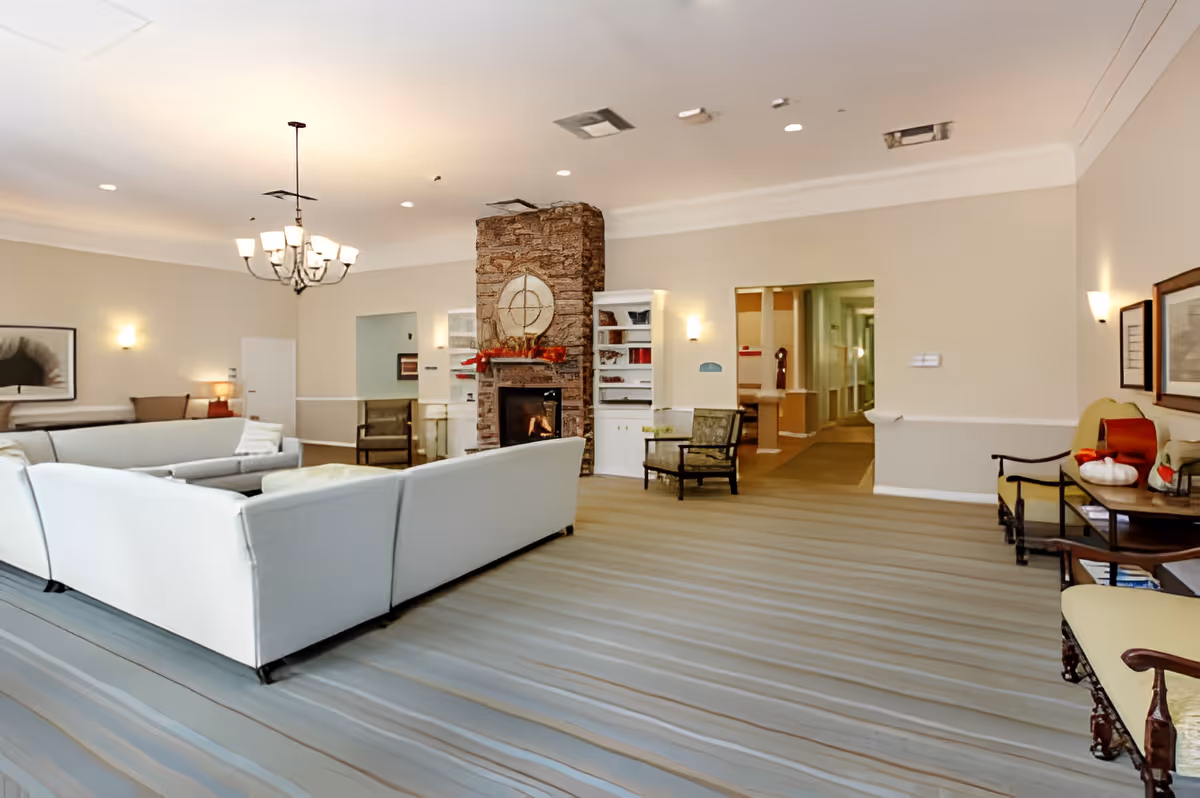 Spacious common lounge with white sectional sofas arranged around a stone fireplace, chandelier, and additional seating in a bright room.