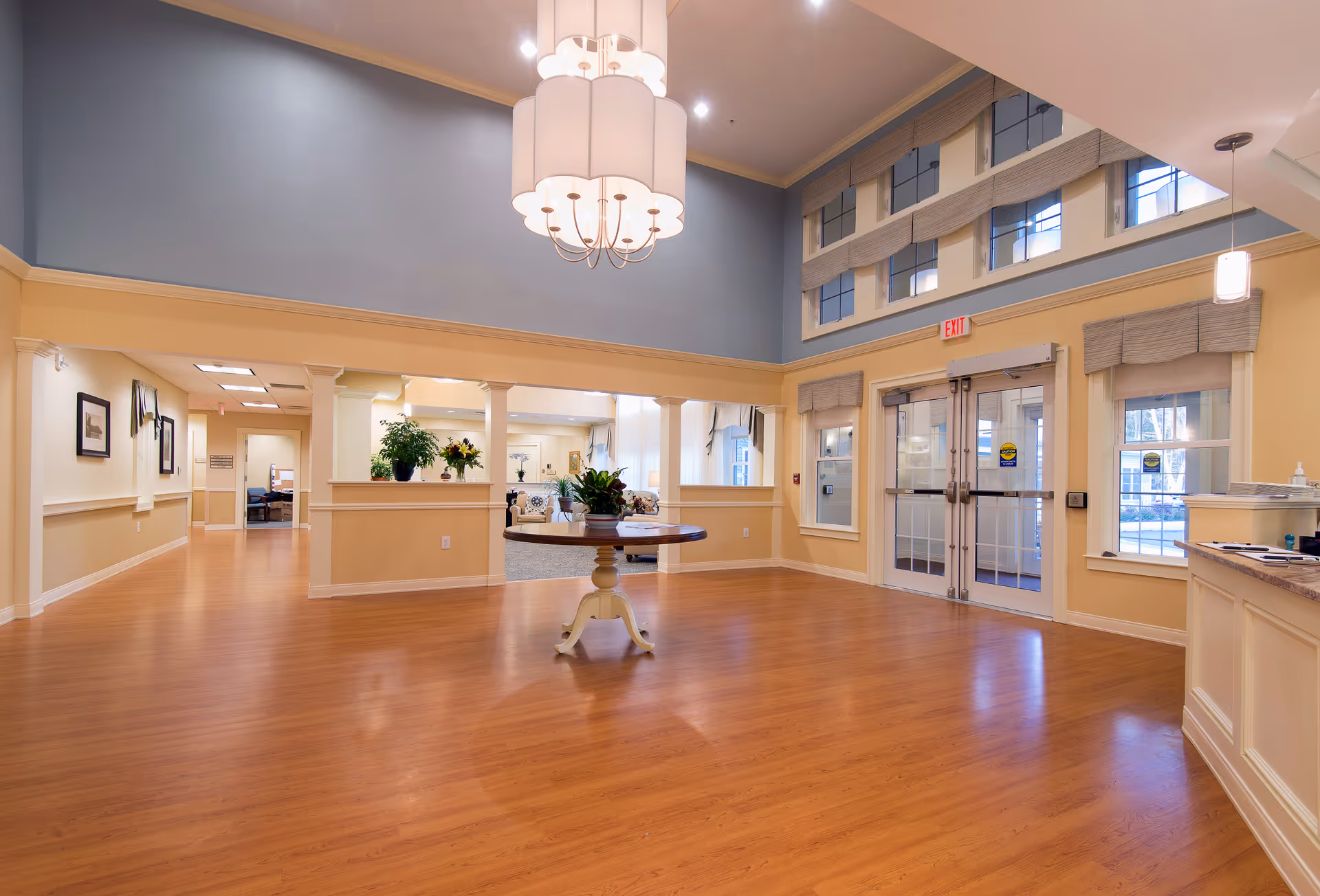 Spacious and well-lit interior lobby area with wooden flooring, a round table with plants in the center, large windows with blinds, double glass doors, and a high ceiling with a modern chandelier. The walls are painted in soft yellow and blue tones, and there is a reception desk on the right side.