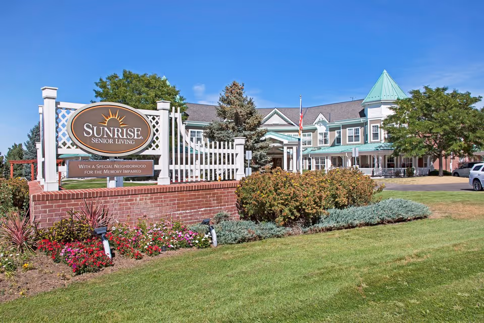 Exterior view of Sunrise Senior Living facility with a large sign in front that reads 'Sunrise Senior Living - With a Special Neighborhood for the Memory Impaired'. The building has a green roof, white trim, and a turret, surrounded by well-maintained landscaping and a clear blue sky.