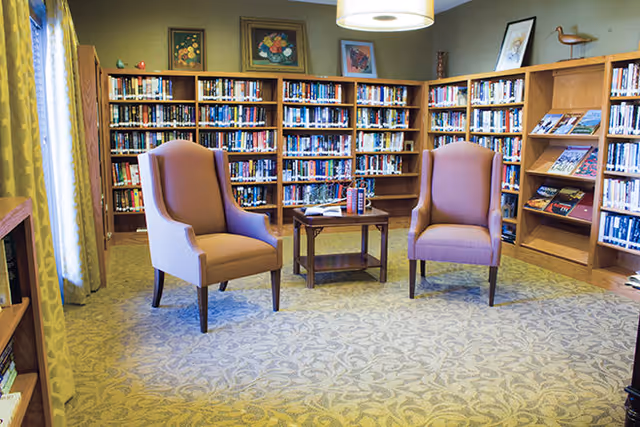 A cozy library room with two upholstered armchairs facing a small wooden table. The walls are lined with wooden bookshelves filled with books, and there are framed pictures and decorative items on top of the shelves. A large window with curtains allows natural light to enter the room.