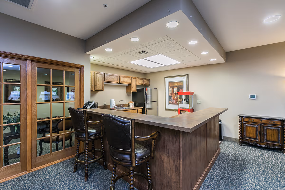 Interior view of a common area with a kitchen and bar counter. The kitchen features wooden cabinets, a stainless steel refrigerator, a sink, and a popcorn machine. Two leather bar stools are positioned at the counter. There are glass-paneled wooden doors on the left and a framed picture on the wall behind the counter. The room has carpeted flooring and recessed ceiling lights.