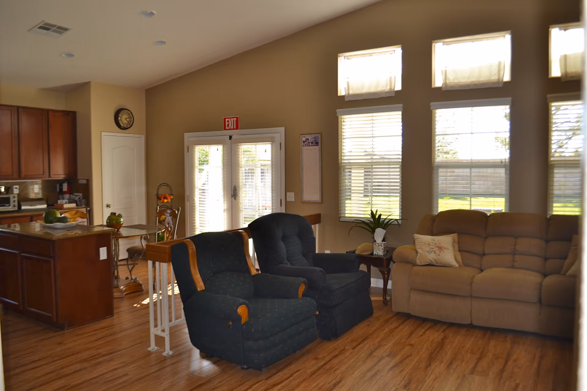 A cozy senior living room area with two dark upholstered armchairs and a beige sofa with a decorative pillow. The room has large windows with blinds allowing natural light to enter. To the left, there is a kitchen area with wooden cabinets, a small dining table with chairs, and a clock on the wall. The floor is wooden, and there is an exit door with an illuminated red exit sign above it.