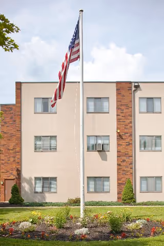 Exterior view of a three-story building with beige and brick facade, several windows, and an American flag on a flagpole in front, surrounded by a flower bed and green lawn under a partly cloudy sky.