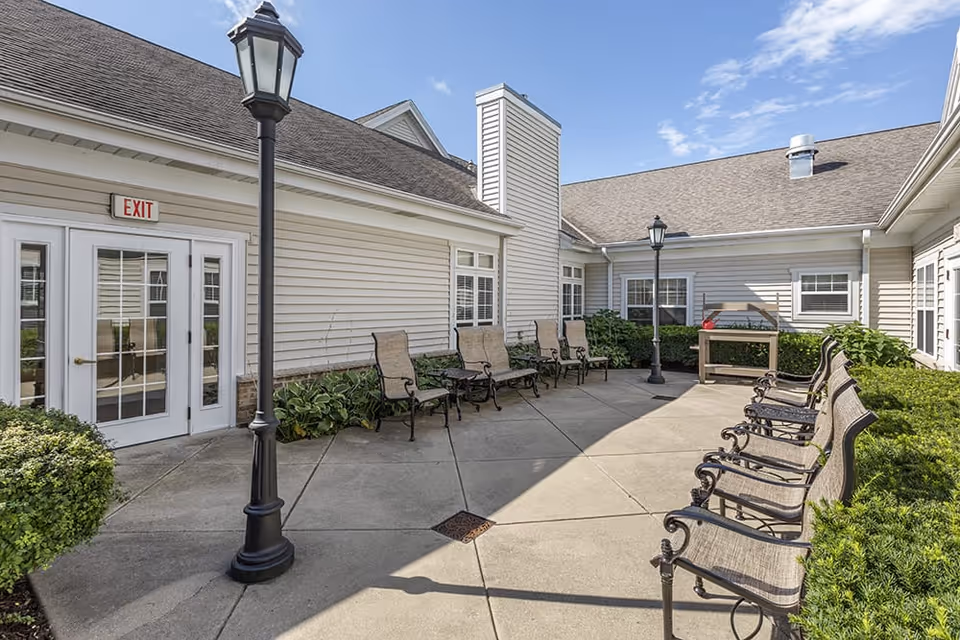 Outdoor courtyard area at Brookdale Kenosha featuring multiple metal and mesh chairs arranged along the walls, two black lampposts, green shrubbery, and beige siding on the surrounding building under a partly cloudy sky.