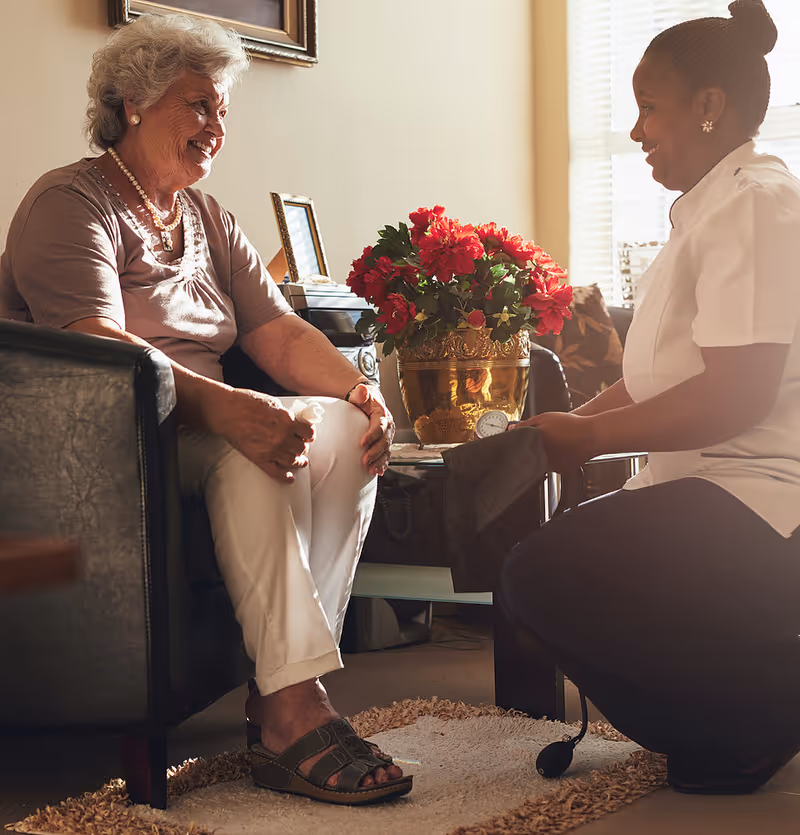 An elderly woman sitting in a chair smiling and interacting with a caregiver who is kneeling in front of her, holding a blood pressure cuff. A vase with red flowers is on a table between them in a warmly lit room.