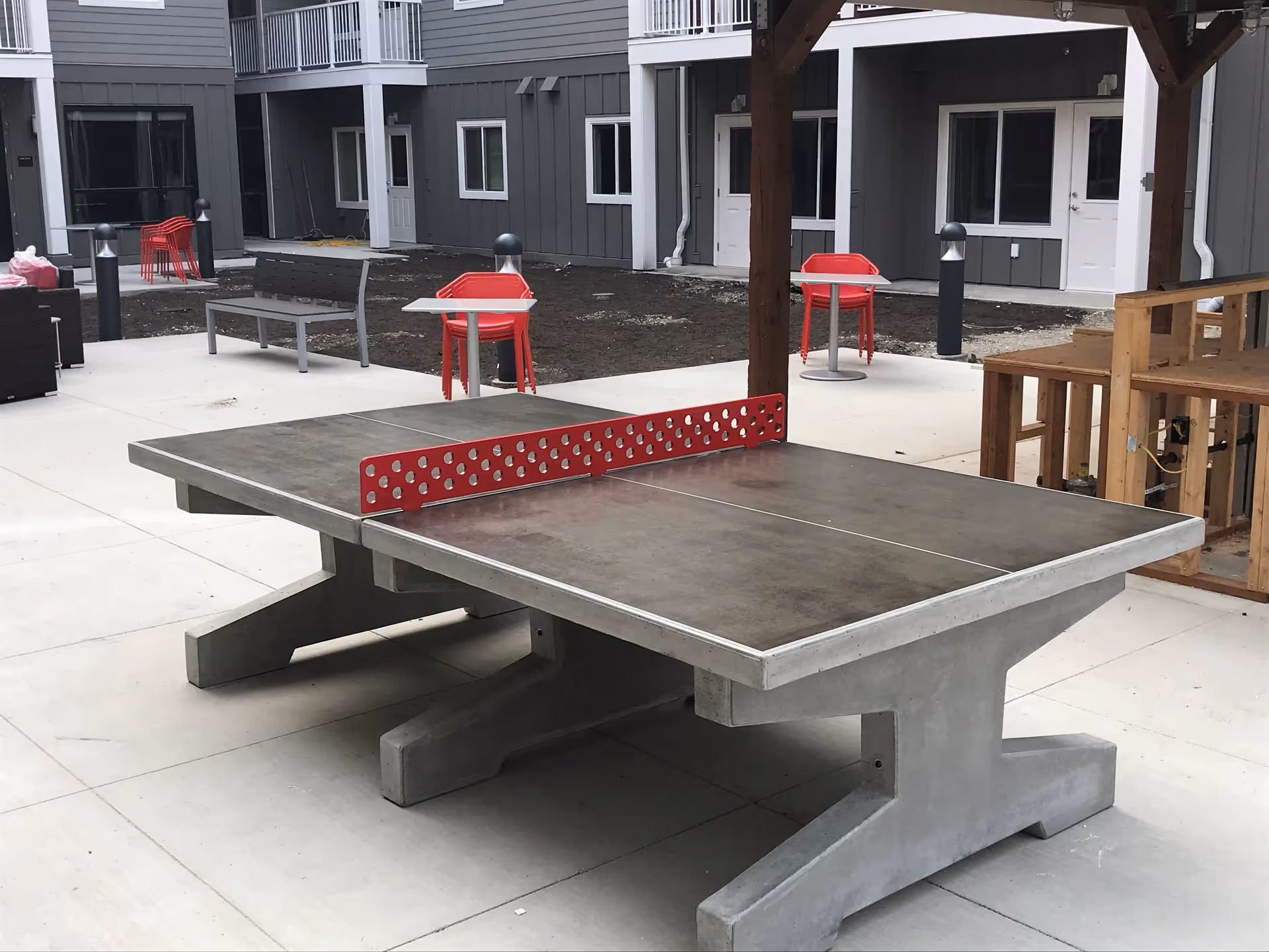 Outdoor area with a concrete ping pong table featuring a red net. Surrounding the table are red chairs, a bench, and a wooden structure. The background shows a gray building with white doors and windows.