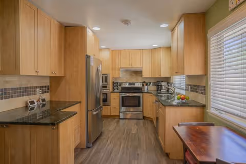 A modern kitchen with light wood cabinets, stainless steel appliances including a refrigerator, oven, and microwave. The countertops are dark granite, and there is a tiled backsplash with a mosaic accent. A wooden table with chairs is positioned near a window with white blinds, allowing natural light to fill the space.