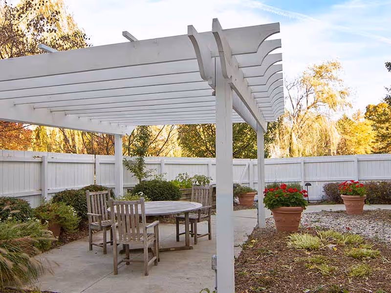 Outdoor patio area with a white pergola overhead, a round wooden table surrounded by four wooden chairs, potted plants with red flowers, and a white fence enclosing the space. Trees with autumn foliage are visible in the background.