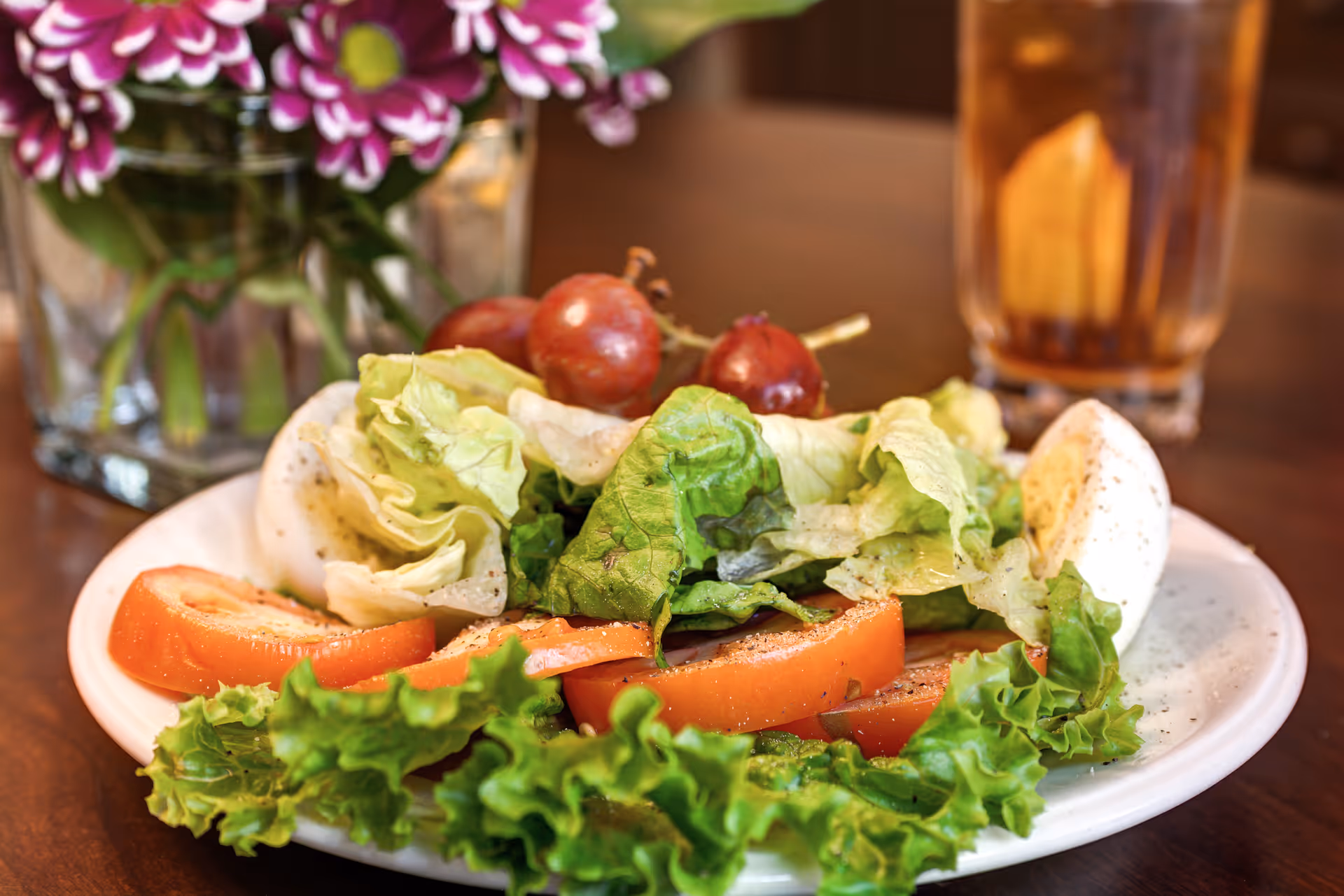 A plate of salad with lettuce, tomato, boiled egg and grapes on a wooden table next to a glass and a vase of flowers.