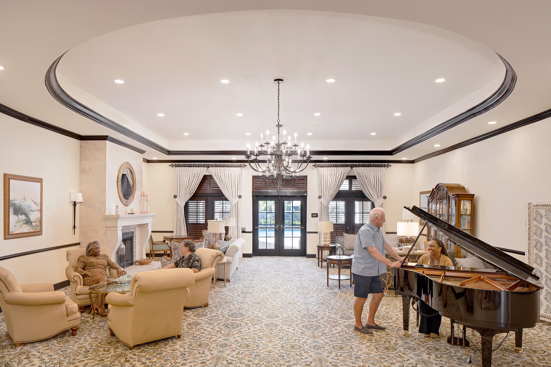 A spacious and elegant living room in a senior living facility with a patterned carpet, beige armchairs arranged around a glass coffee table, and a grand piano on the right side. Two women are seated and conversing near the fireplace on the left, while a man stands next to a woman playing the piano on the right. The room features large windows with curtains, a chandelier hanging from the ceiling, and a door leading outside in the background.