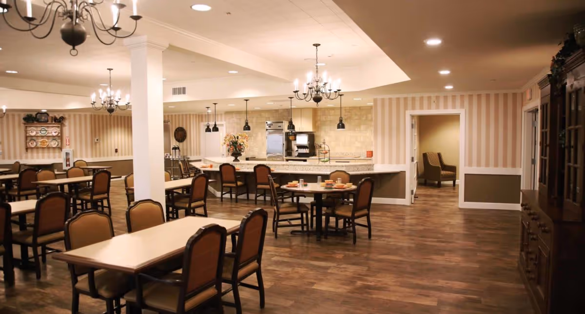 A spacious dining room in a memory care community with multiple tables and chairs arranged neatly. The room features wood flooring, striped wallpaper, chandeliers, and a kitchen area with a counter and bar stools in the background. There is a doorway leading to another room with an armchair visible.