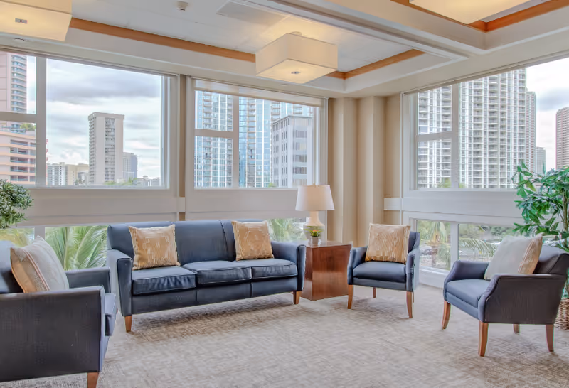 A bright and modern living room area with large windows showing a cityscape view. The room features a dark blue sofa and three matching armchairs, each with beige patterned throw pillows. There is a wooden side table with a lamp and a small plant, and another larger plant is placed near the window. The ceiling has recessed lighting and a square light fixture.