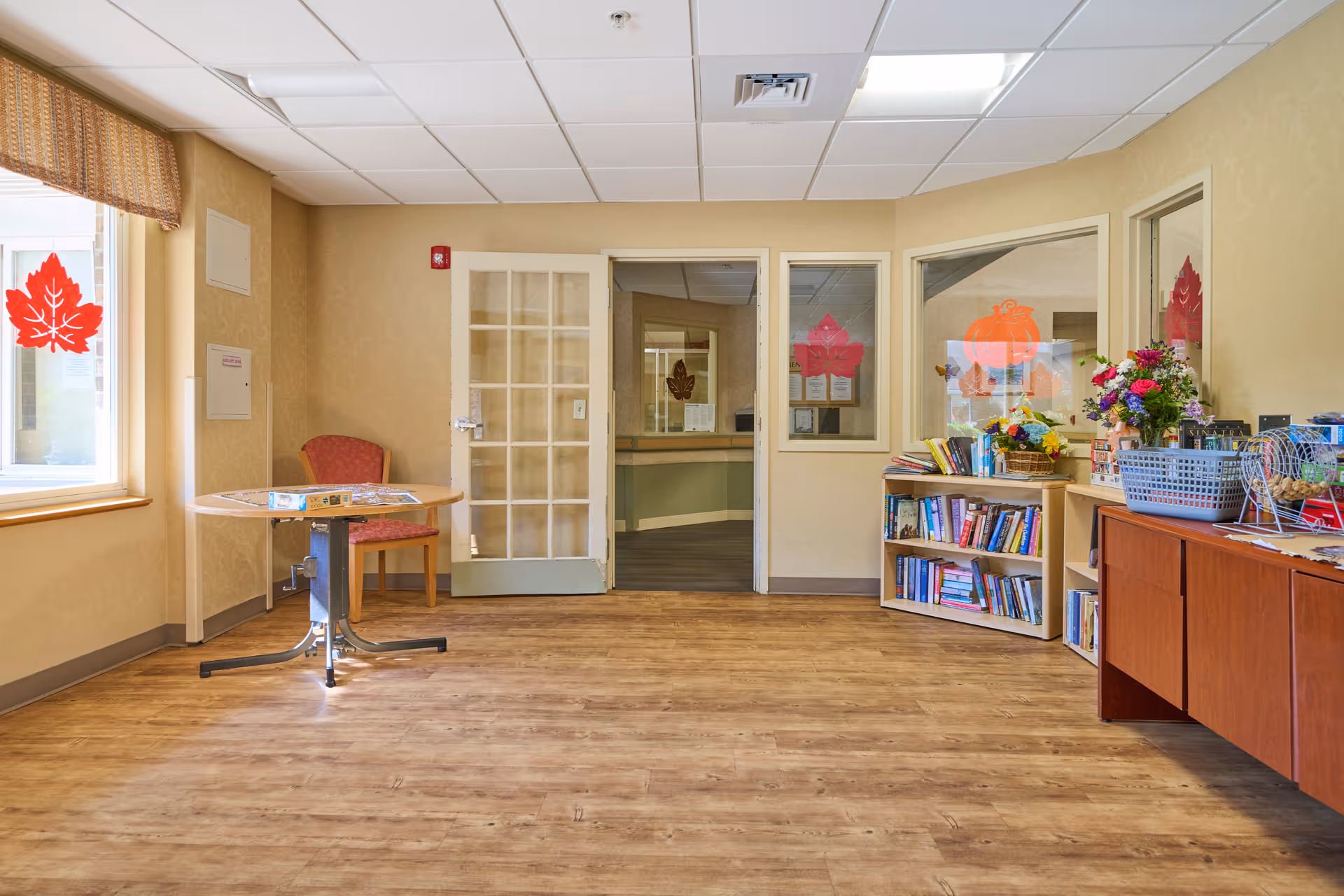 A bright room with wooden flooring and beige walls featuring a round table with a red chair near a window decorated with a red maple leaf decal. On the right side, there is a bookshelf filled with books, a basket of flowers, and a cabinet with various items. The room has a door with glass panes and two windows with autumn-themed decals, including a pumpkin and maple leaves.