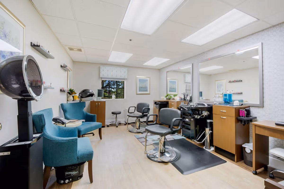 Interior of a hair salon area in a senior living facility with two teal armchairs, two black salon chairs in front of large mirrors, hair drying machines, and wooden cabinets. The room has light-colored walls, a window with a view of trees, and ceiling lights.