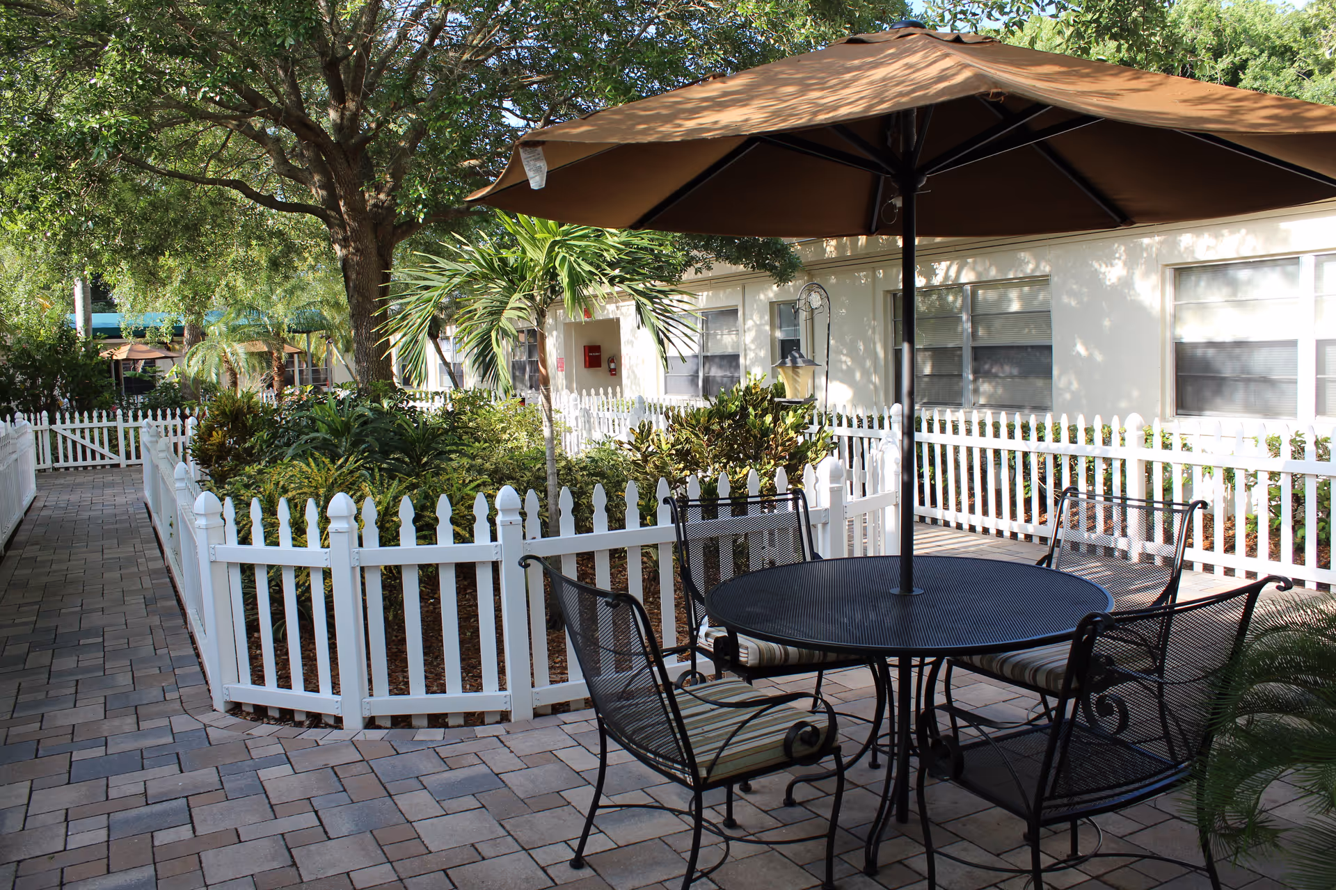Shaded outdoor courtyard with a round metal table and chairs under a large umbrella next to a white picket fence and landscaped garden.