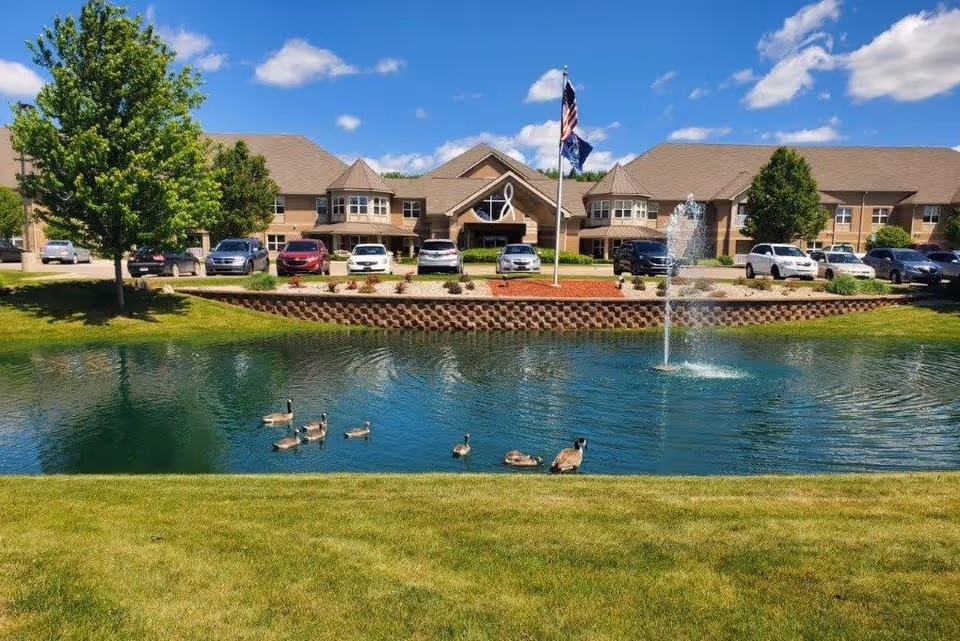 Exterior view of Village Manor Retirement facility with a pond in the foreground containing several ducks, a water fountain in the pond, a grassy area, trees, parked cars, and a building under a blue sky with scattered clouds.
