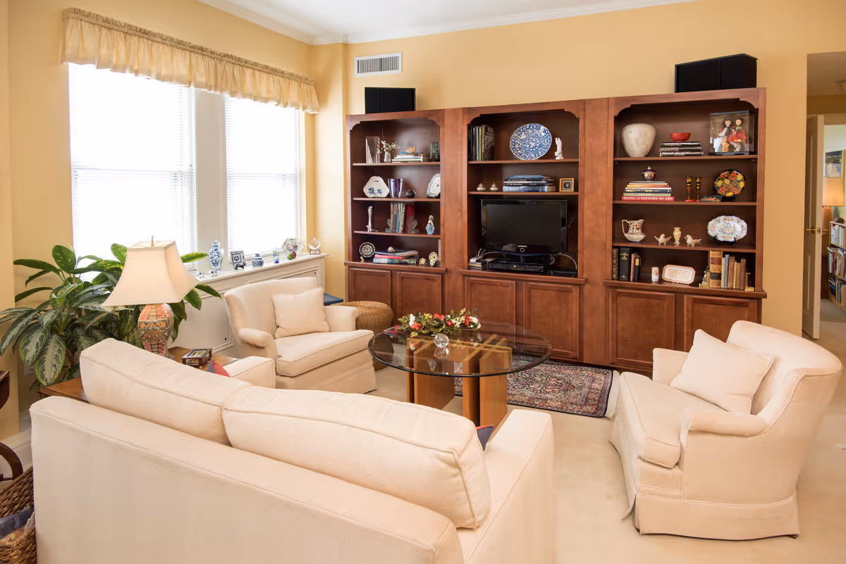 A cozy living room with beige upholstered sofa and armchairs arranged around a glass coffee table. Behind the seating area is a large wooden entertainment center with shelves displaying decorative plates, books, and various ornaments. A window with blinds and a valance lets in natural light, and a table lamp and green plants add warmth to the space.