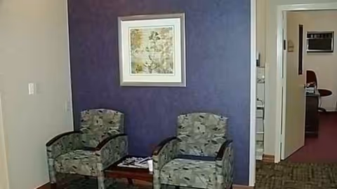 Two patterned chairs with a small table sit beside a purple wall and framed art.