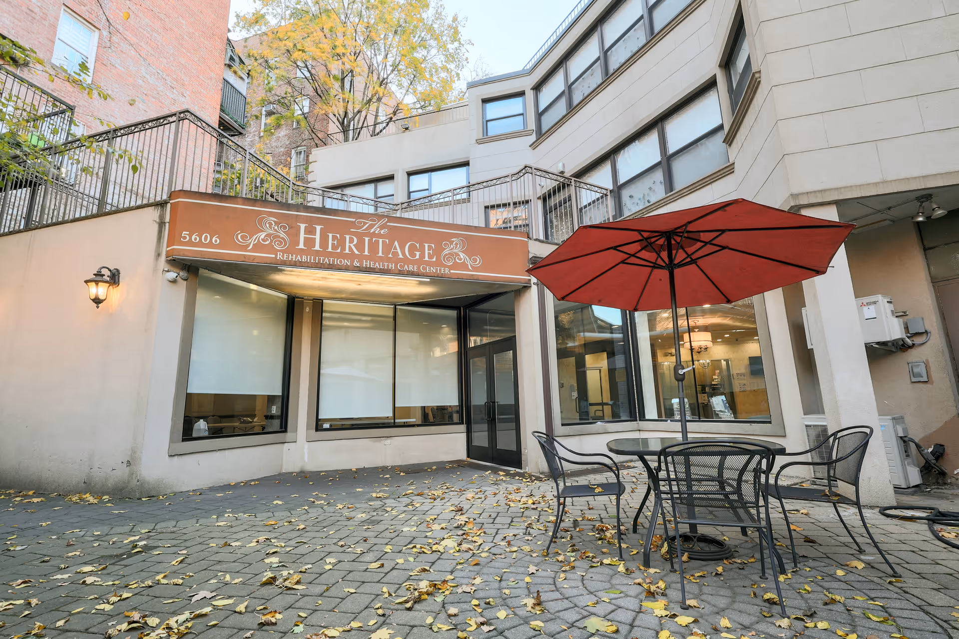 Outdoor patio area of The Heritage Rehabilitation & Health Care Center with a round metal table, four chairs, and a large red umbrella. The building entrance and windows are visible, along with fallen leaves scattered on the paved ground.