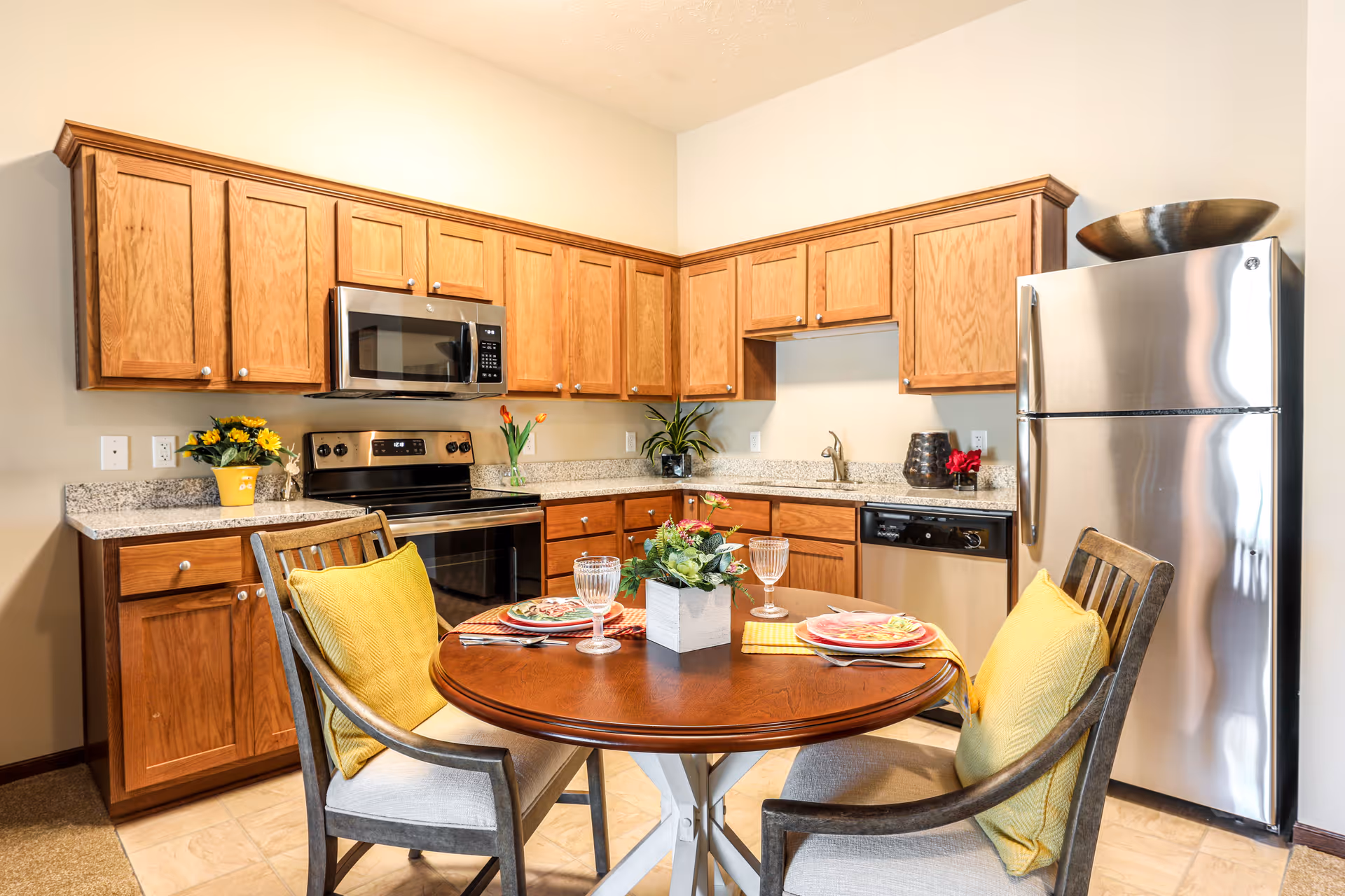 A bright kitchen with wooden cabinets, stainless steel appliances including a microwave, stove, dishwasher, and refrigerator. In the foreground, there is a round wooden dining table set for two with plates, glasses, and silverware, and two chairs with yellow cushions. The countertop has decorative plants and flowers.