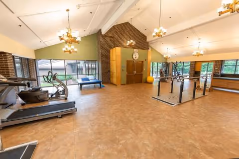A spacious senior living fitness room with exercise equipment including a treadmill, elliptical machine, and weightlifting area. The room has large windows letting in natural light, brick walls, and multiple ceiling lights.