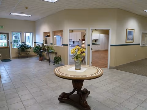 Interior view of a spacious assisted living facility lobby with tiled flooring, a round wooden table with a vase of yellow flowers in the center, potted plants near windows and doors, and doorways leading to other rooms including a kitchen area.