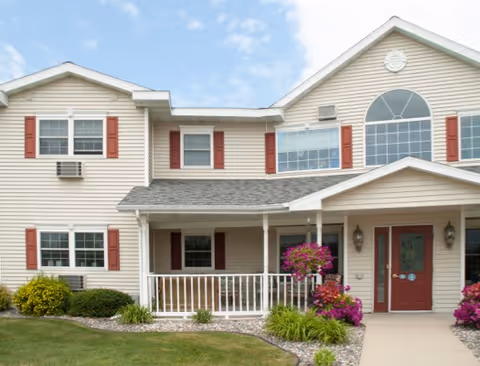 Exterior front view of a two-story residential building with beige siding, red shutters, and a covered porch entrance. There are well-maintained shrubs and flowering plants along the walkway leading to the red front door under a clear blue sky.