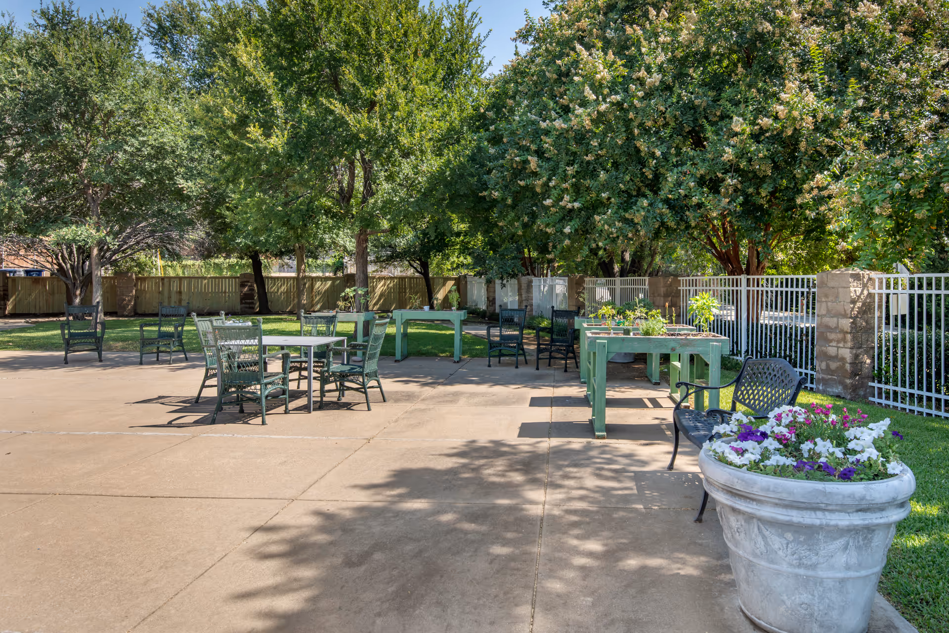 Outdoor patio area with several green metal chairs and tables arranged on a concrete surface. There are green trees and plants surrounding the area, a white metal fence along the right side, and a large planter with purple and white flowers in the foreground.