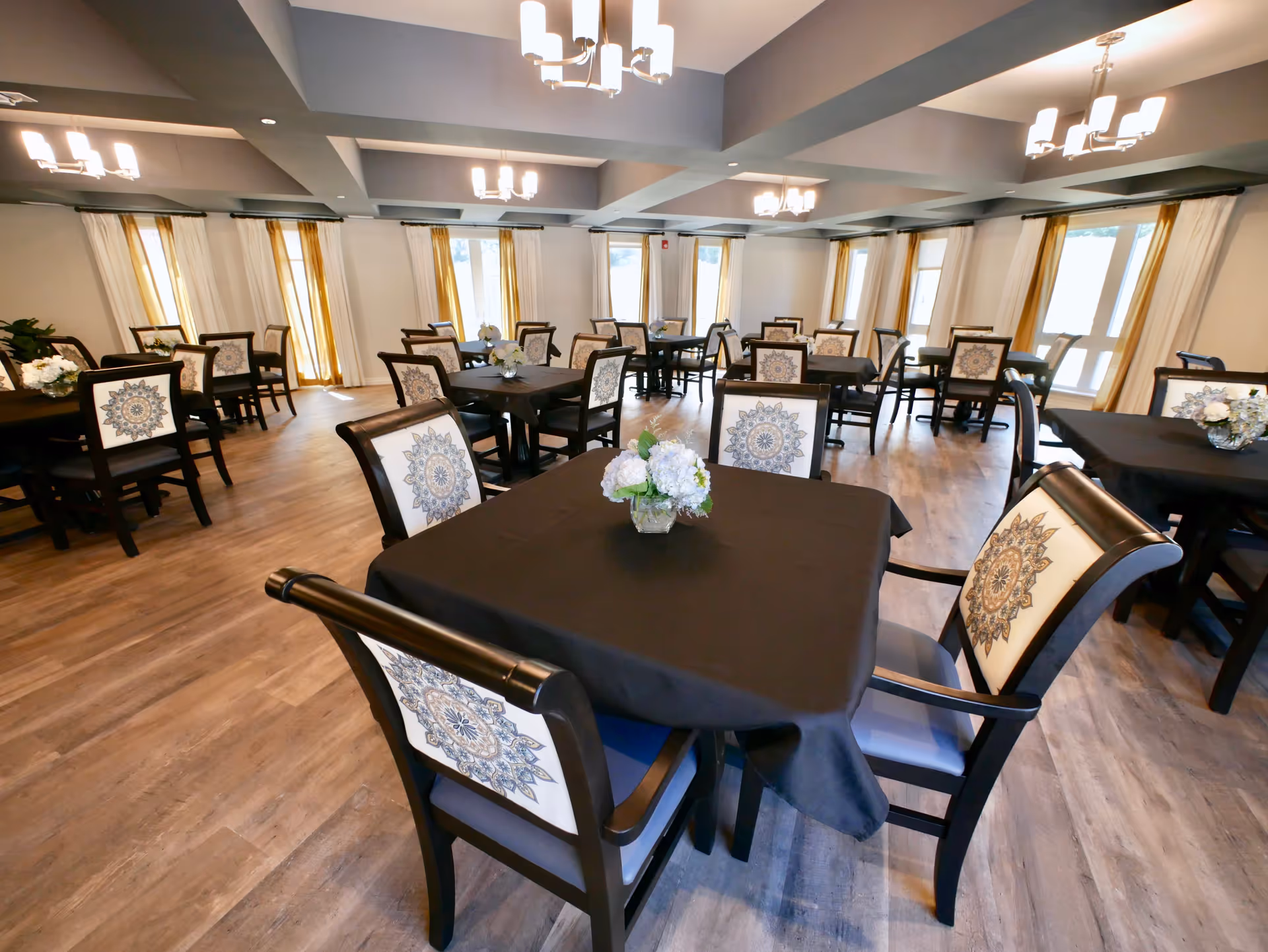 A spacious dining room with multiple square tables covered in black tablecloths, each surrounded by four chairs with decorative patterned backs. The room features large windows with white and yellow curtains, wooden flooring, and modern ceiling lights.
