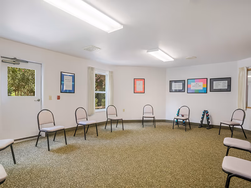 A spacious room with a circle of chairs arranged around the perimeter. The room has beige carpet, white walls, and fluorescent ceiling lights. There are windows with beige curtains and framed pictures on the walls. A door with a window shows greenery outside. Two small racks with colorful weights are placed near the wall.