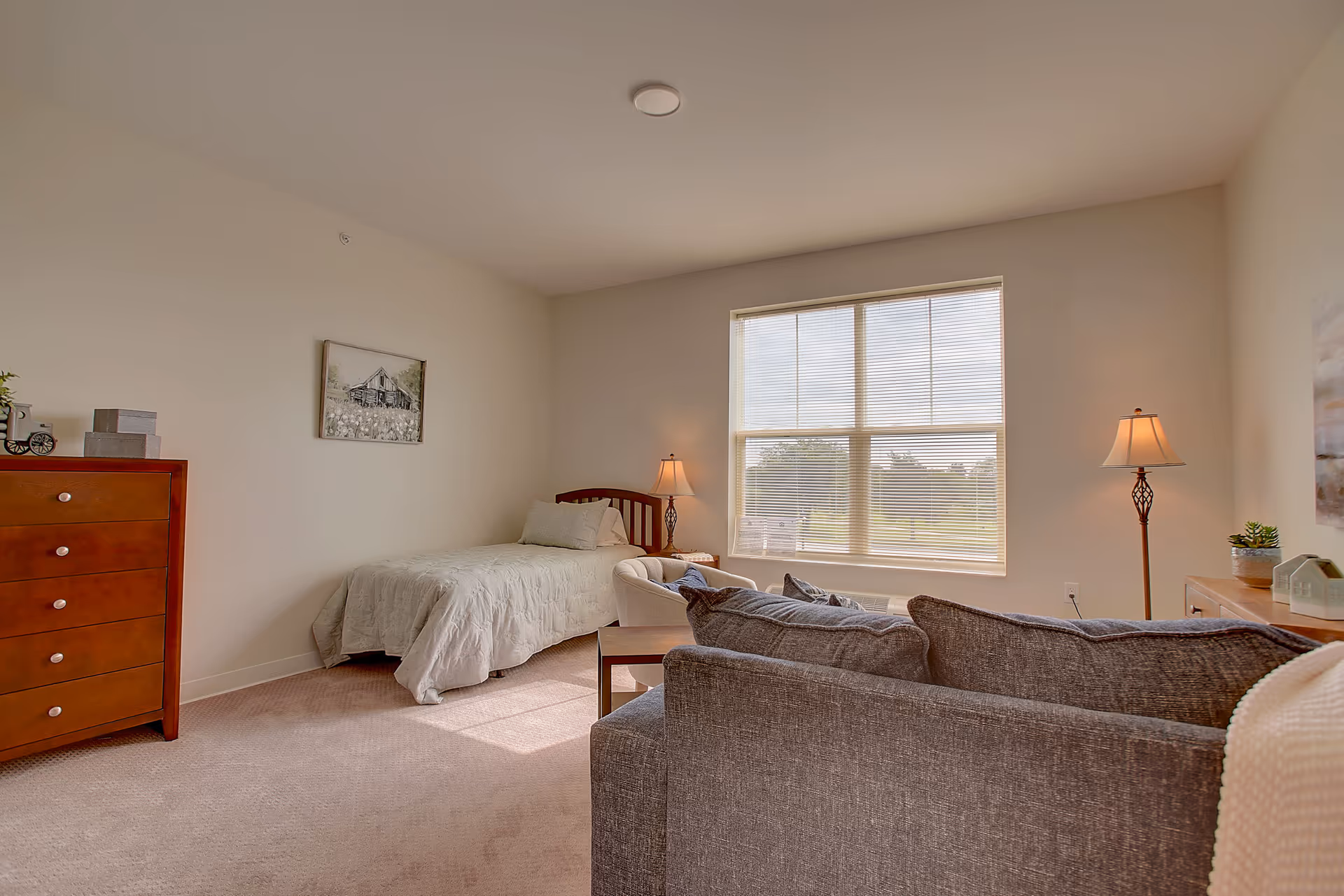 Bright senior living studio room with a bed, sofa, dresser, and a large window.