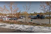 Street view of a single-story nursing facility with a parking lot, sidewalk, and leafless trees with several parked cars.